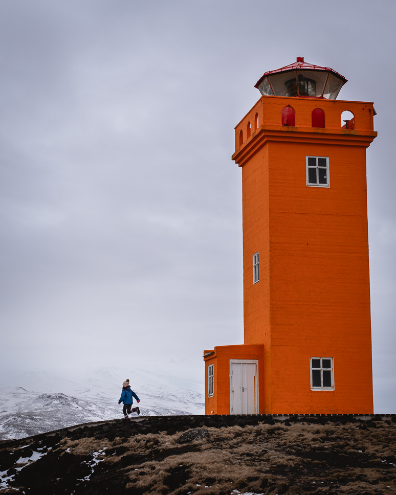 20+ Best things to do in Iceland: Our Ultimate Iceland Bucket List Girl runs away from orange lighthouse with snow-capped mountains in the background on the Snæfellsnes peninsula.