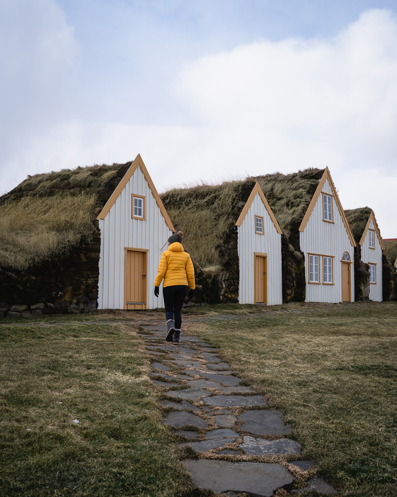 woman with yellow jacket walking to one of the glaumbaer turf houses in iceland