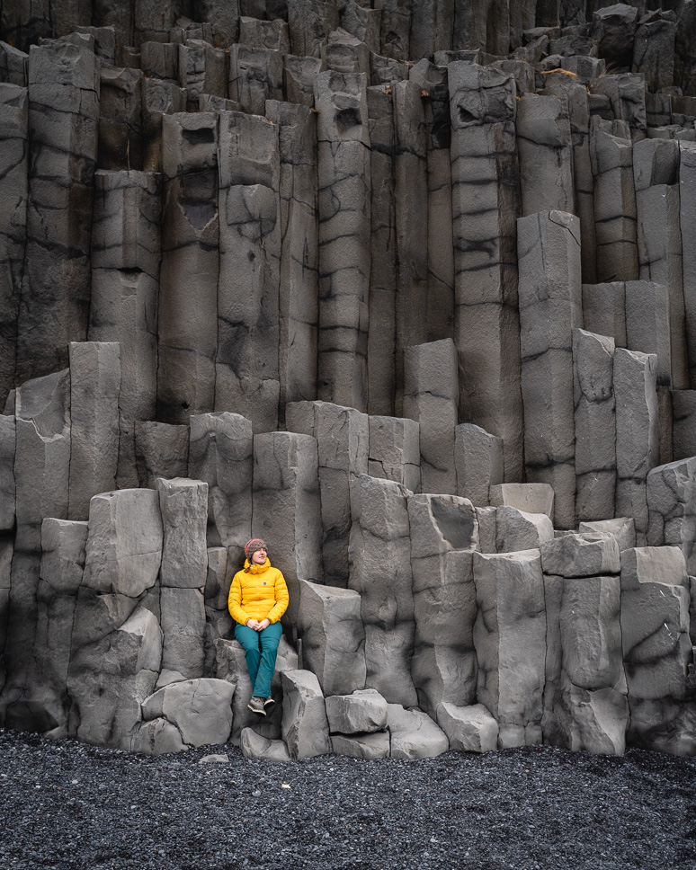 20+ Best things to do in Iceland: Our Ultimate Iceland Bucket List Woman in a yellow jacket sits on the basalt rocks of Reynisfjara Black Beach, a Black sand beach in South Iceland