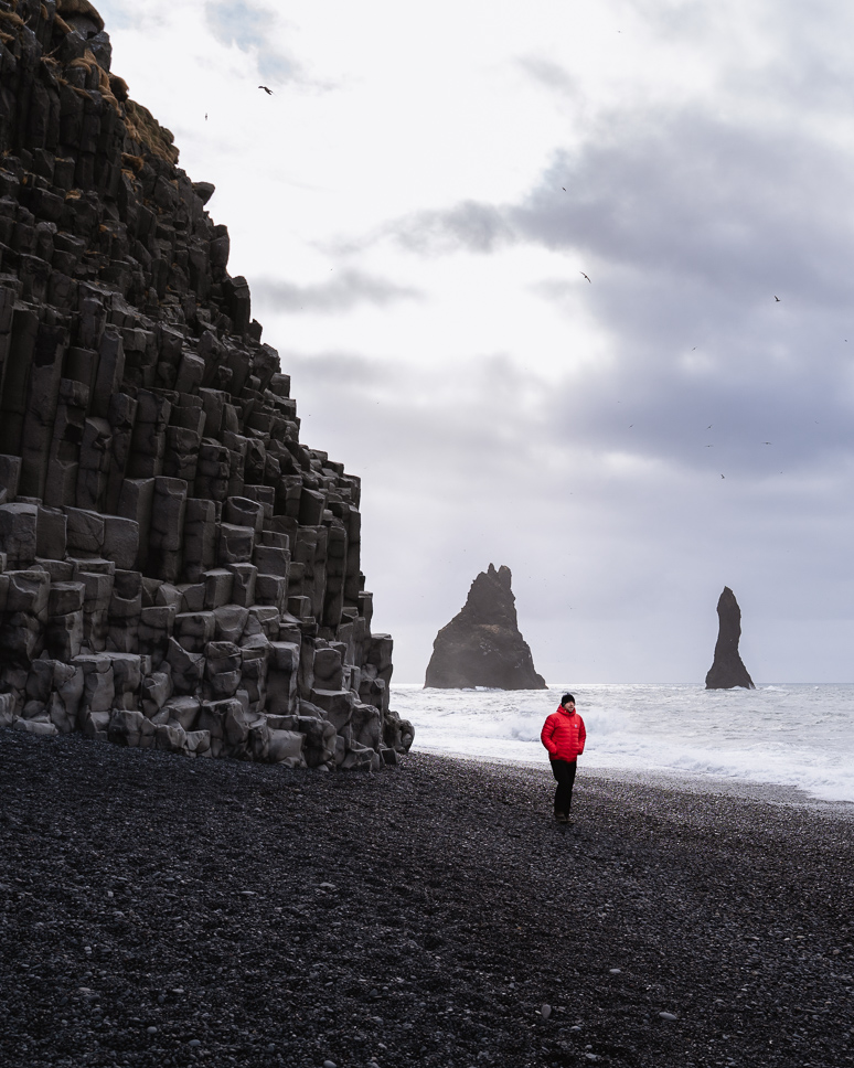 20+ Best things to do in Iceland: Our Ultimate Iceland Bucket List Man in red coat walking on Reynisfjara Black Beach, with the basalt rocks and rock formations in the background